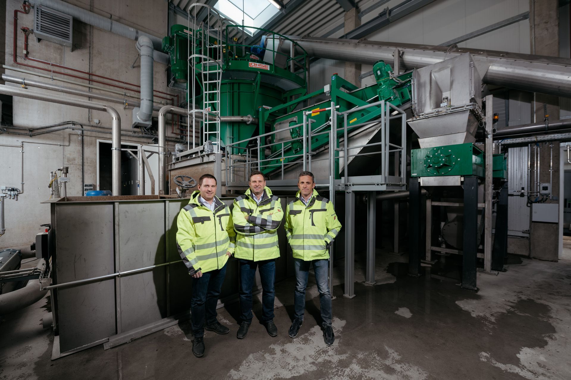 Three workers in safety jackets pose in front of machinery in a facility.