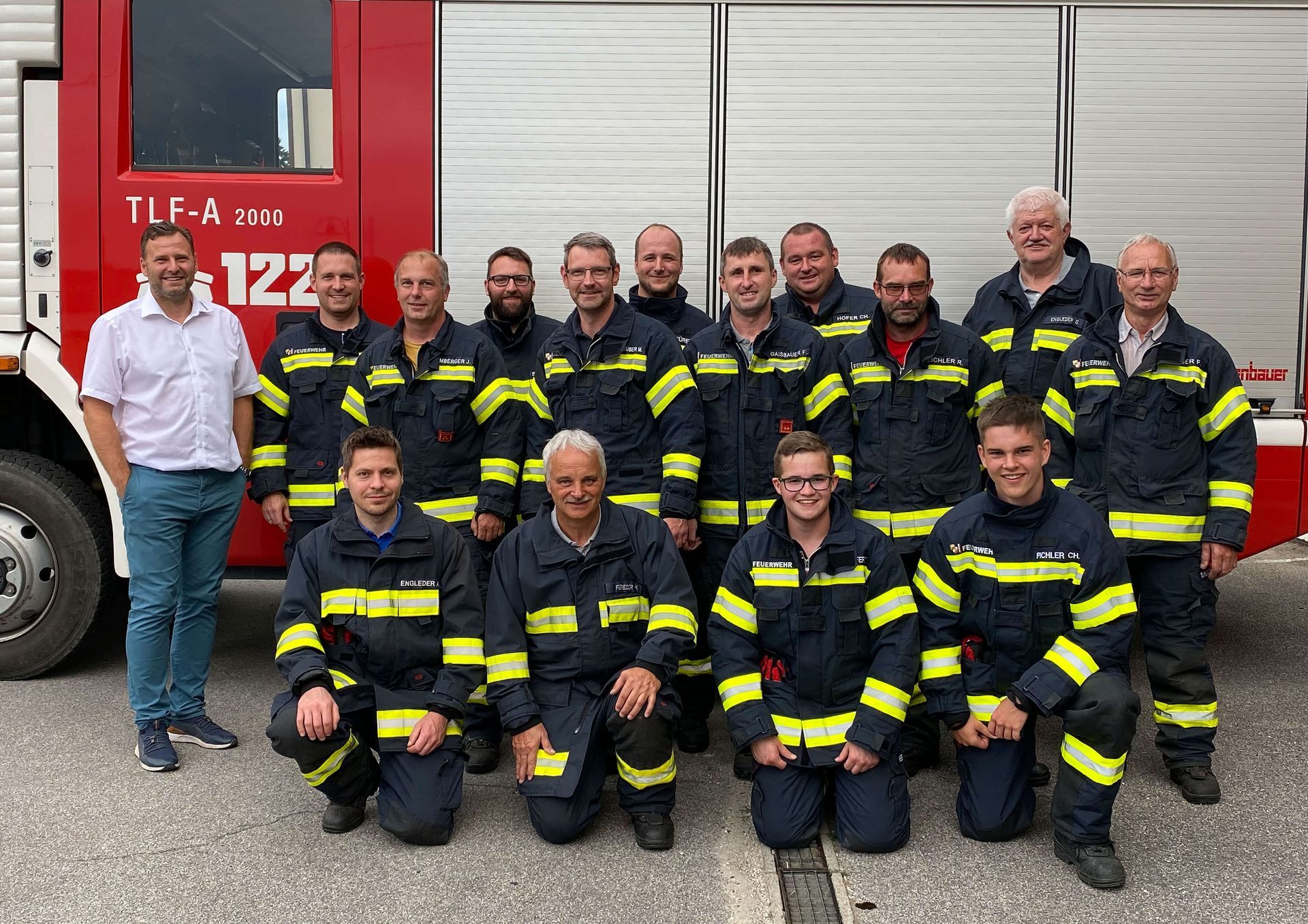 Firefighters in uniform pose in front of a red fire truck; a man in a white shirt stands at the left.