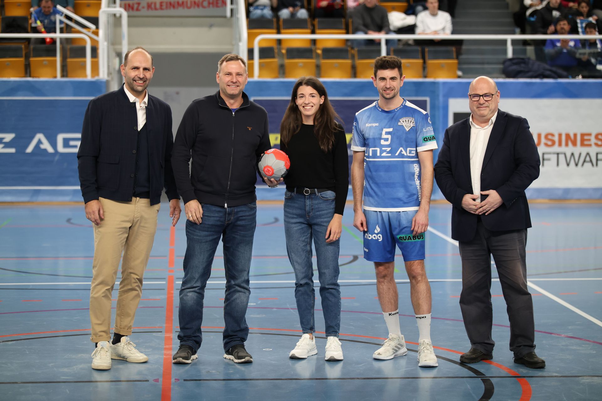 Five people stand on a court, posing for a photo. One holds a handball.