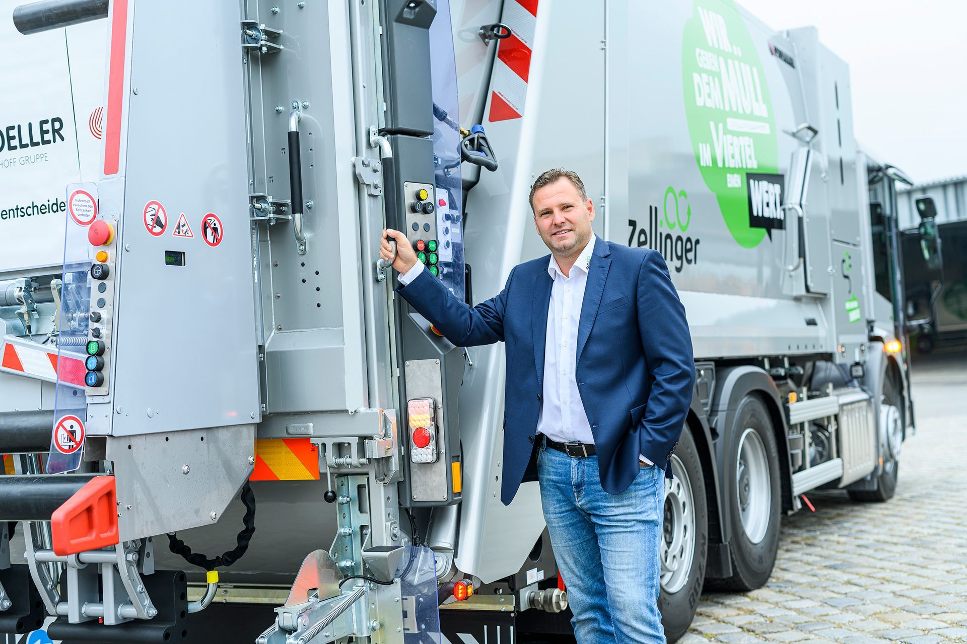 Man in a blue blazer stands next to a waste collection truck. The truck is grey, with green lettering.