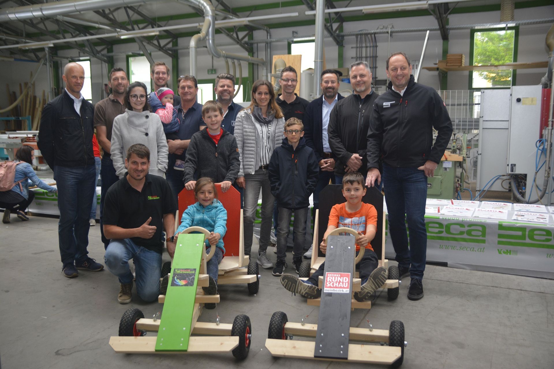 Group of people posing with wooden go-karts in a workshop setting.