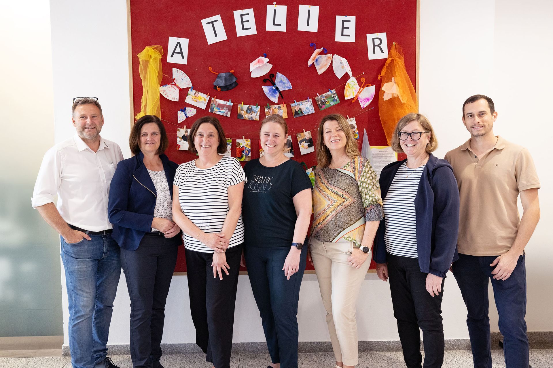 Group of seven people posing in front of a red bulletin board that says