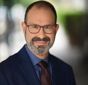 Professional headshot of a smiling man wearing glasses, a blue button-down shirt, a dark tie, and a navy suit.