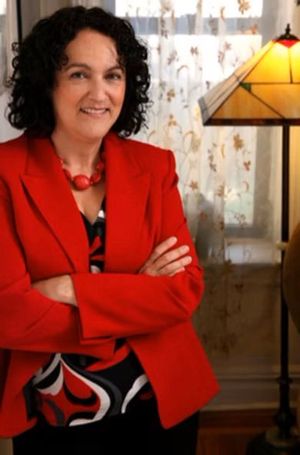 A smiling person with curly dark hair wearing a bright red blazer stands with arms crossed next to a floor lamp.