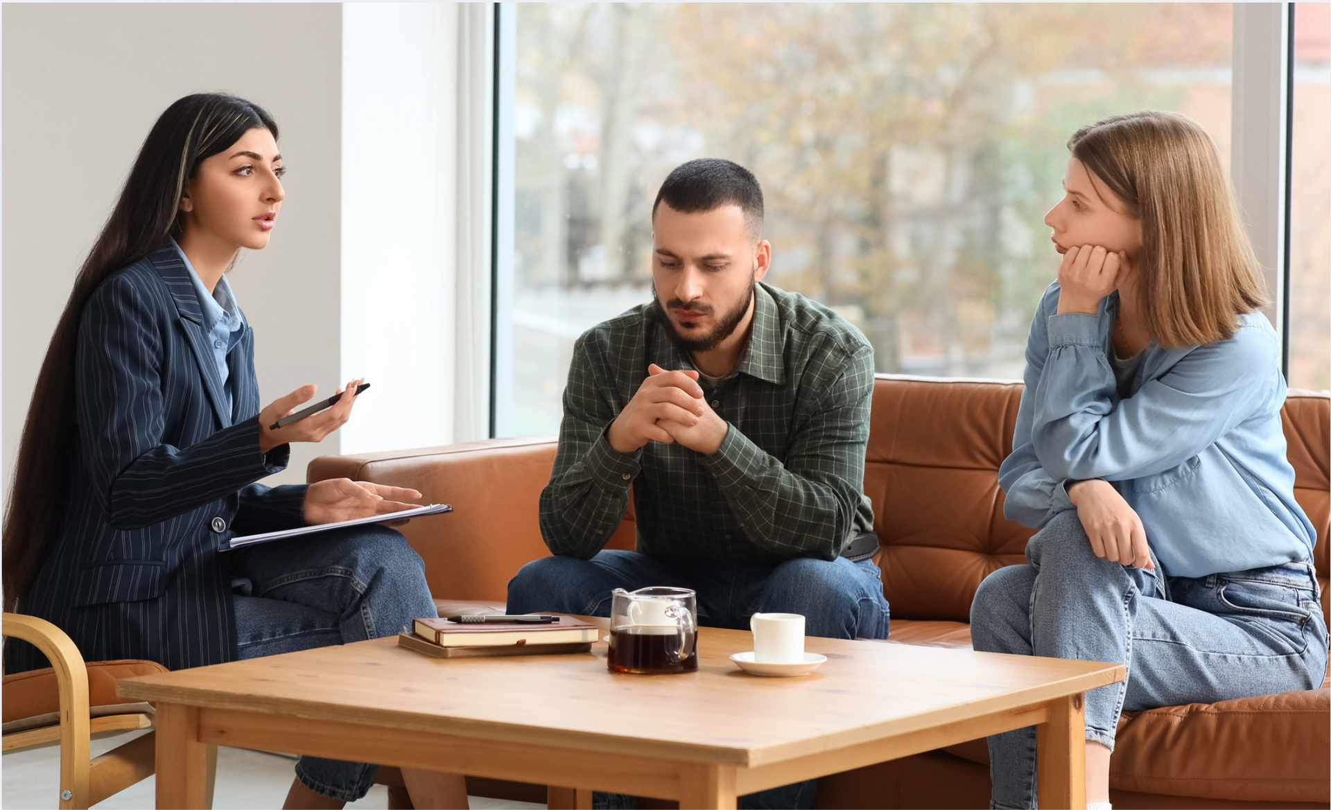 Court mediation meeting with legal documents, gavel, and scales of justice on a wooden desk. A professional sits with a couple in a bright, modern office, discussing documents on a coffee table.