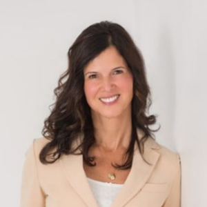 A smiling person with shoulder-length wavy dark hair, wearing a beige blazer over a white shirt, leaning against a wall.