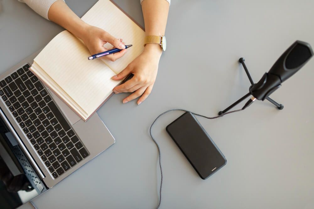 Una persona está escribiendo en un cuaderno al lado de una computadora portátil y un micrófono.