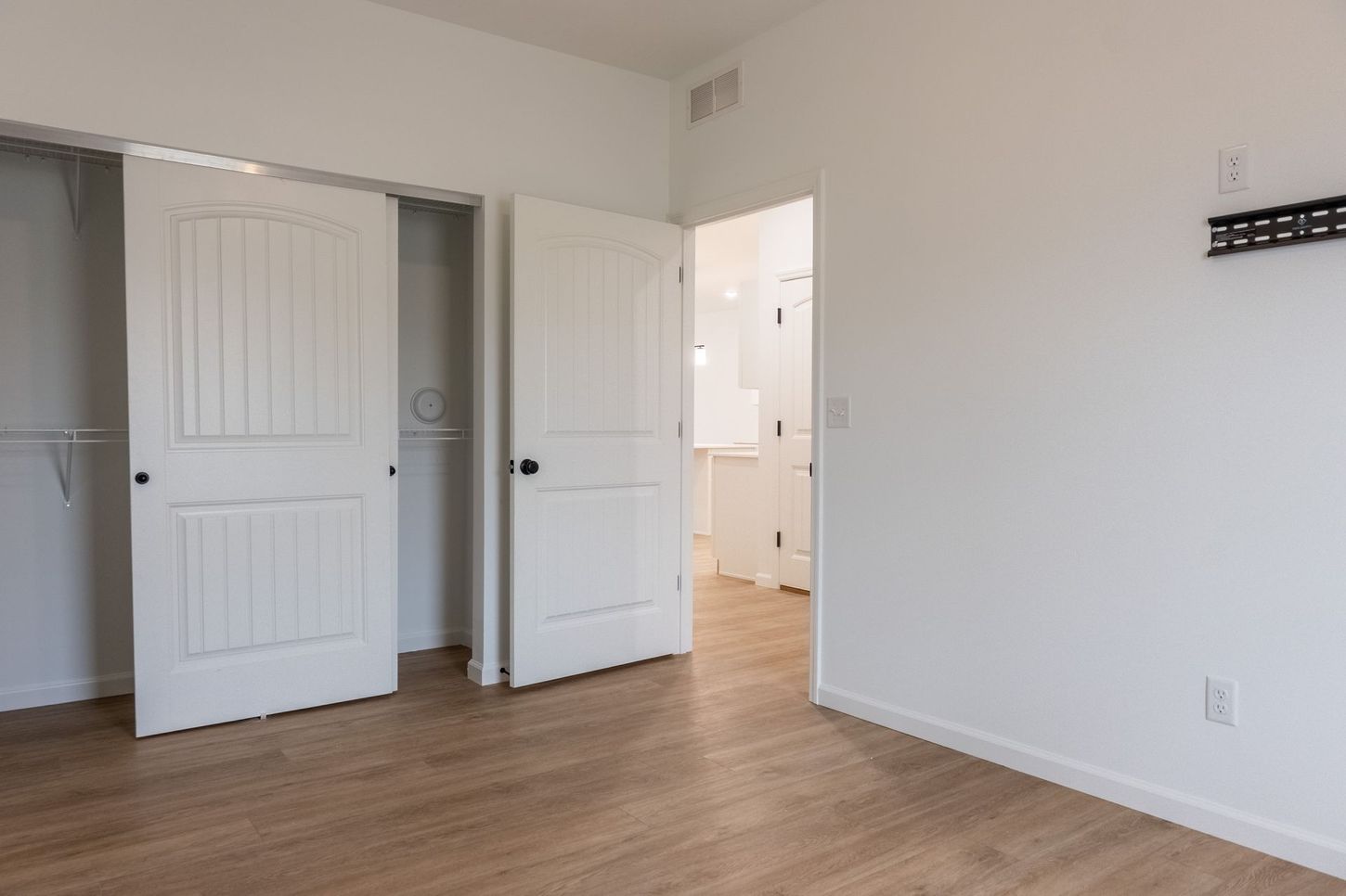 Empty white bedroom with open double closet doors and hardwood floors, leading to a bathroom through an open door