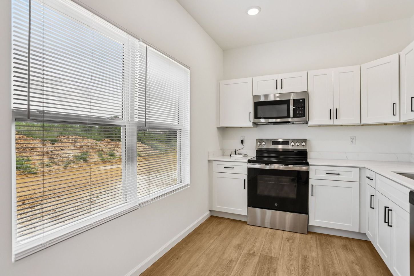 Bright modern kitchen with white cabinets, stainless appliances, wood floor, and large window blinds