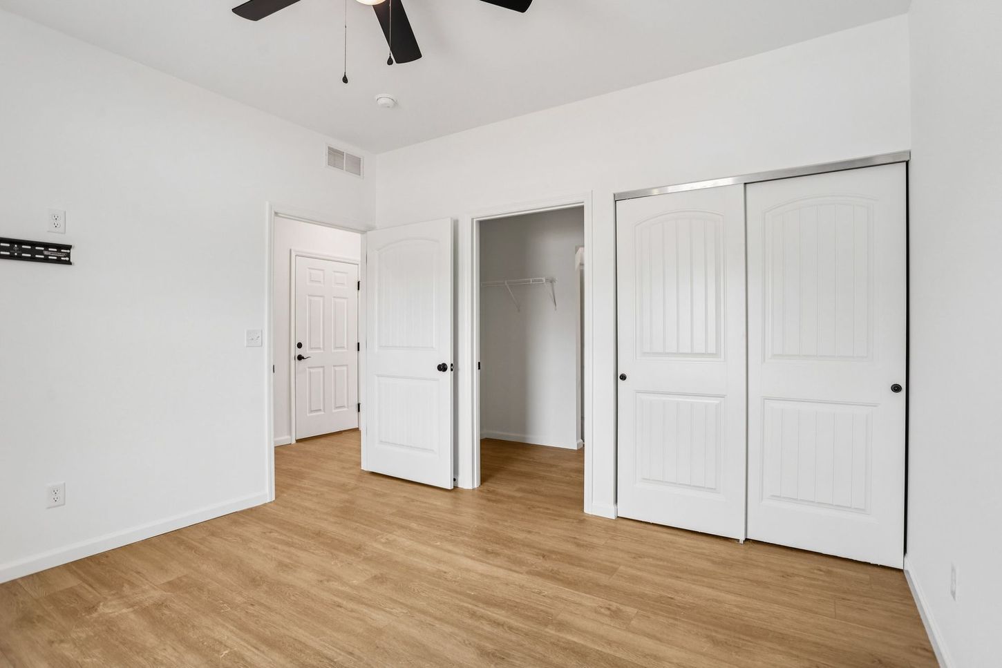 Empty white bedroom with wood flooring, ceiling fan, and open closet doors.