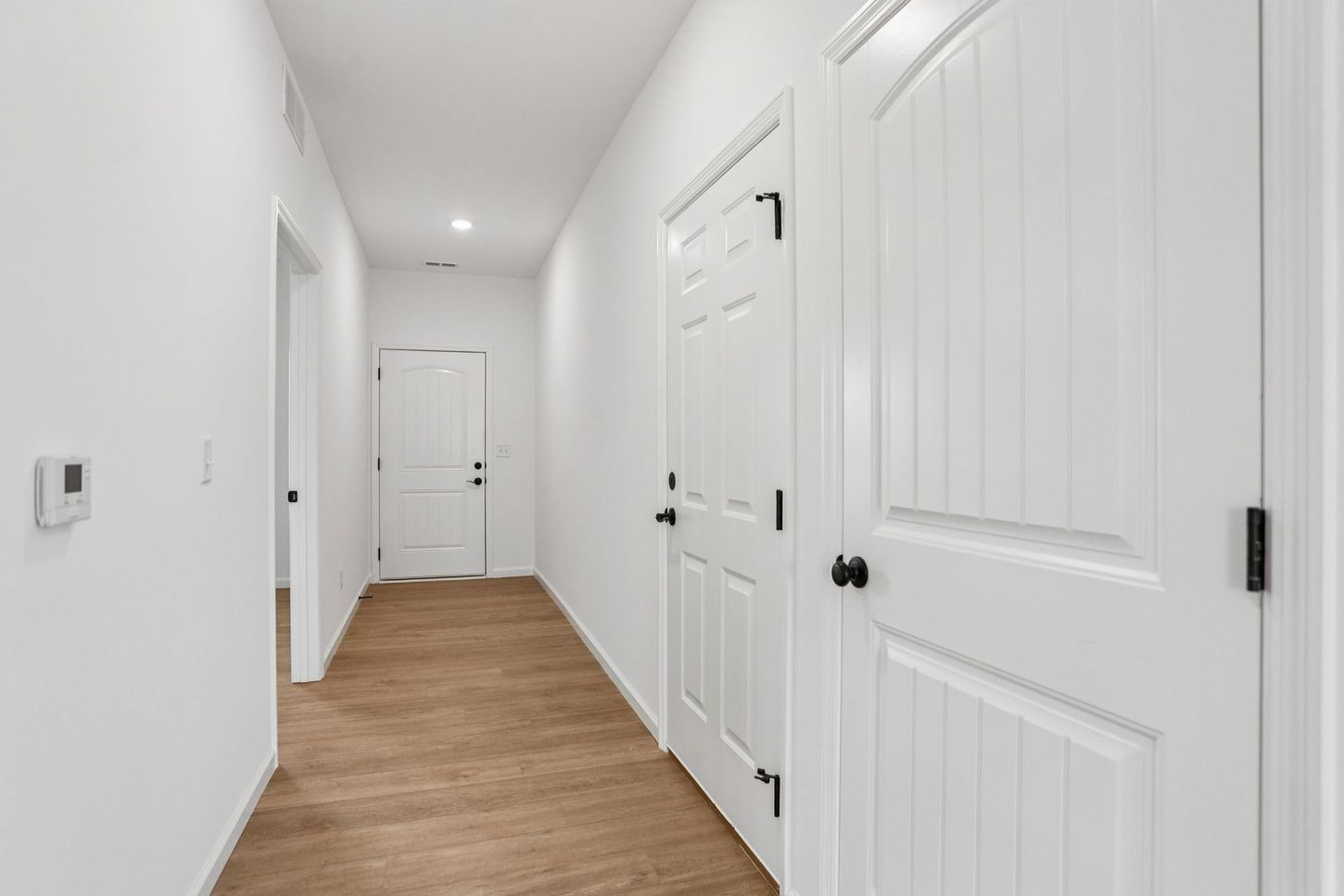 Bright hallway with white doors, white walls, and wood flooring leading to a closed door at the end.