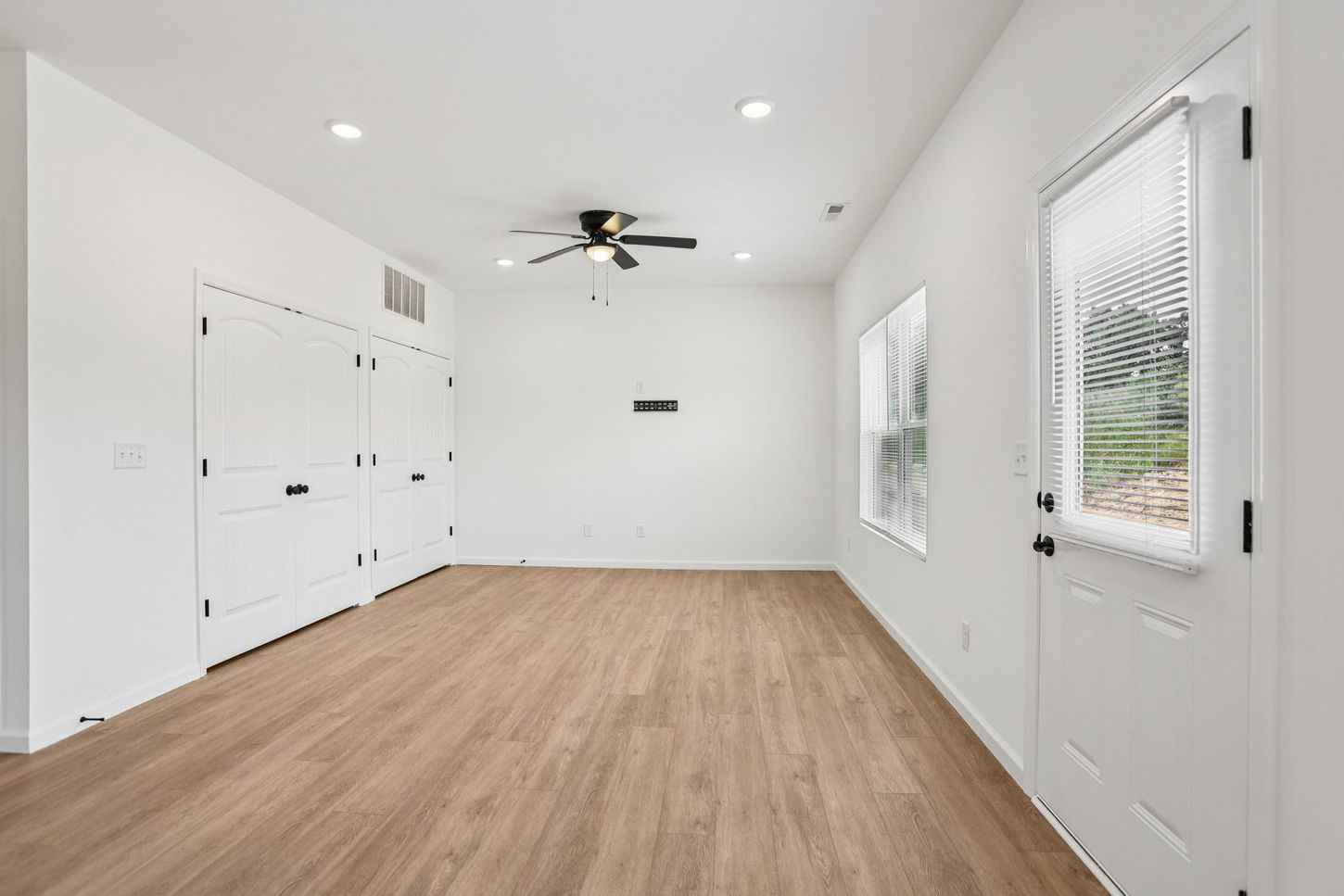 Bright empty room with white walls, wood floors, ceiling fan, closet doors, and windows with blinds