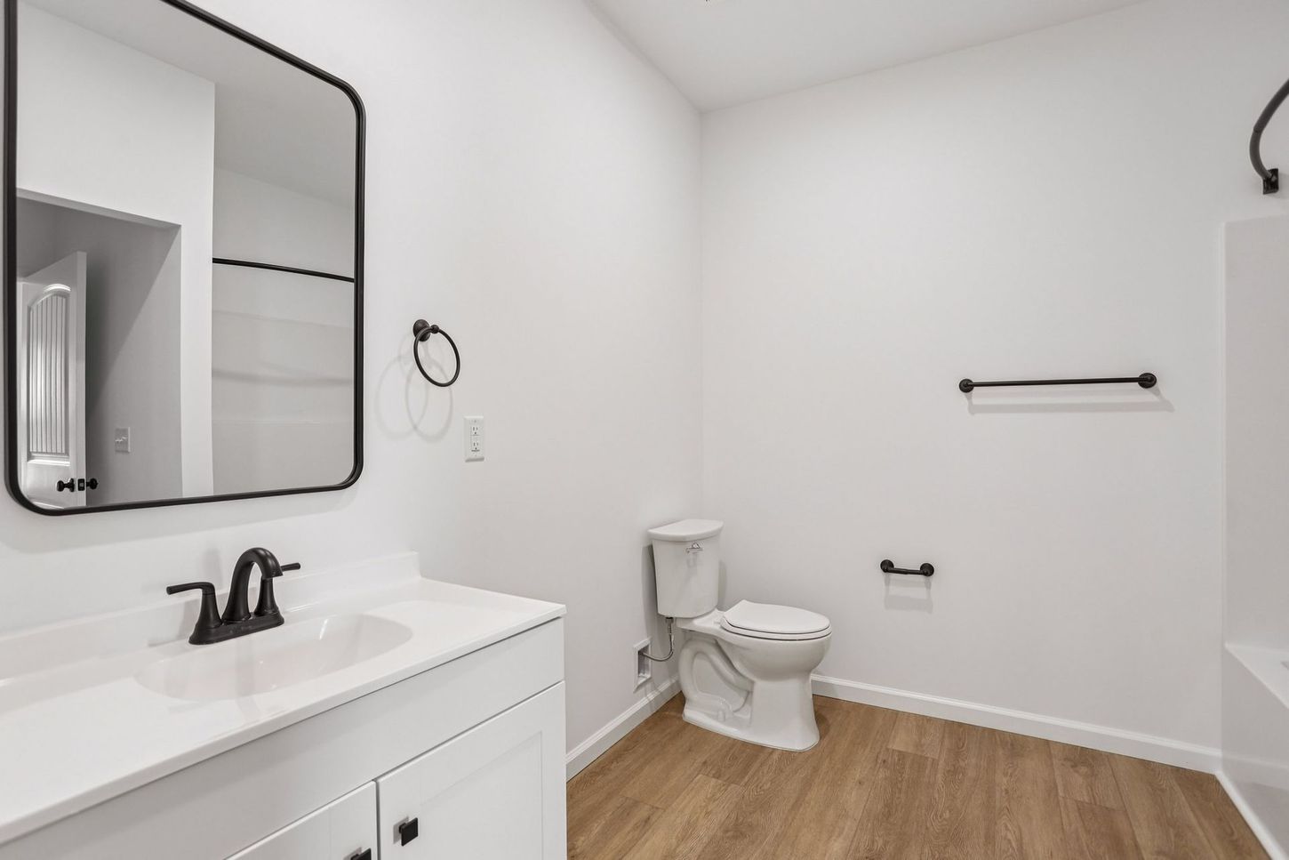 Bright modern bathroom with white vanity, black fixtures, toilet, towel bars, and wood-look floor.