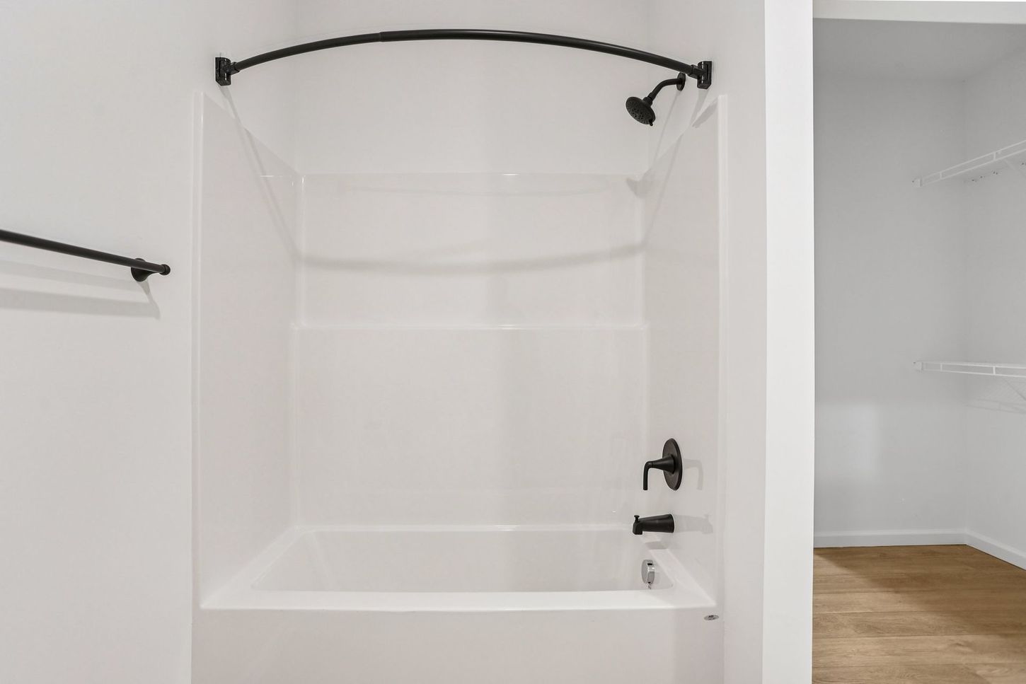 White bathroom with a bathtub, shower rod, and black fixtures beside an open doorway to a wood-floored room