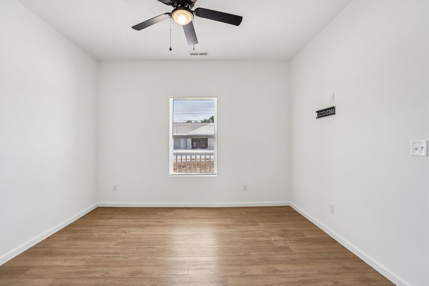 Empty white bedroom with wood floor, ceiling fan, and a single window.