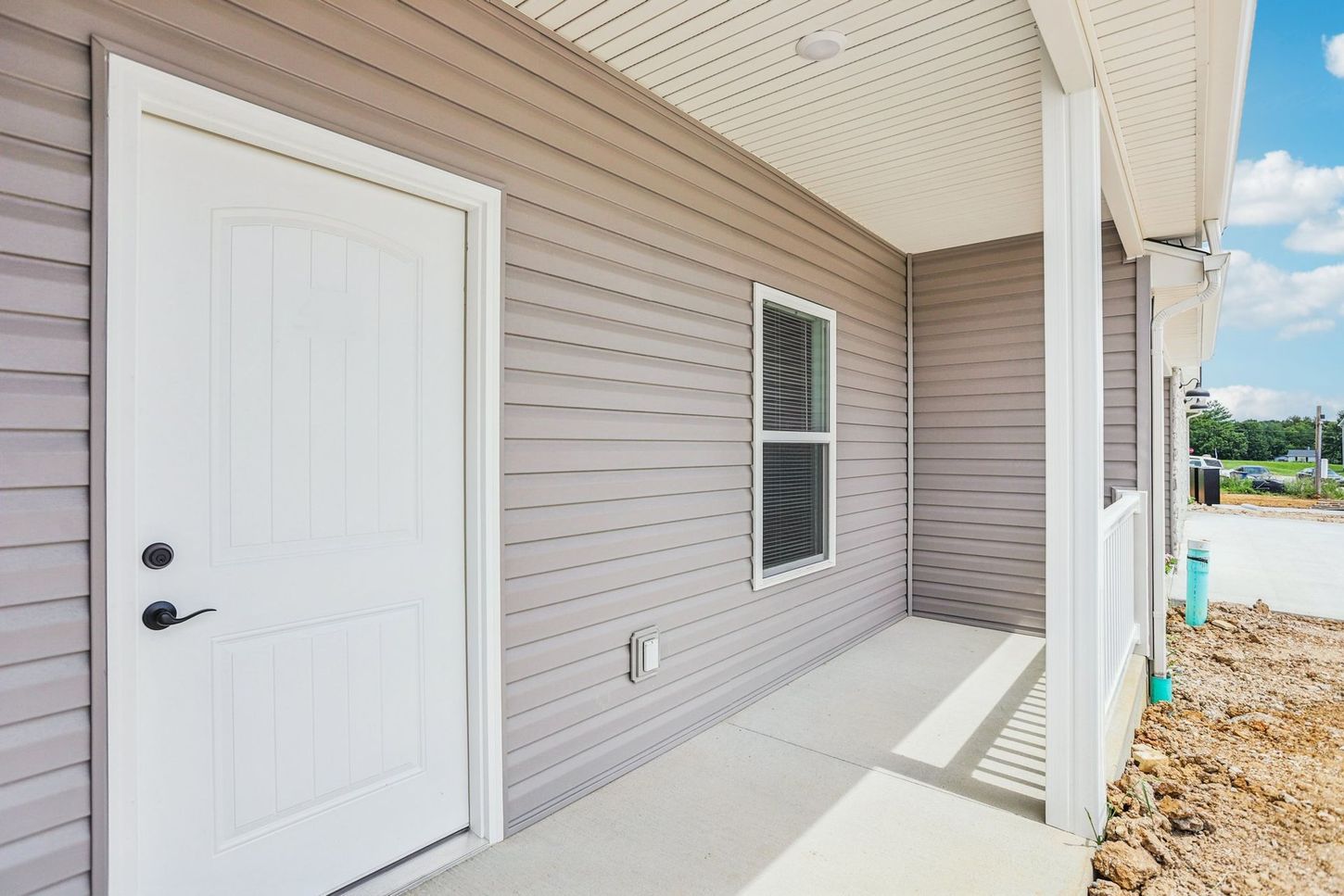 Covered front porch with white door, beige siding, and a small window beside a dirt yard.