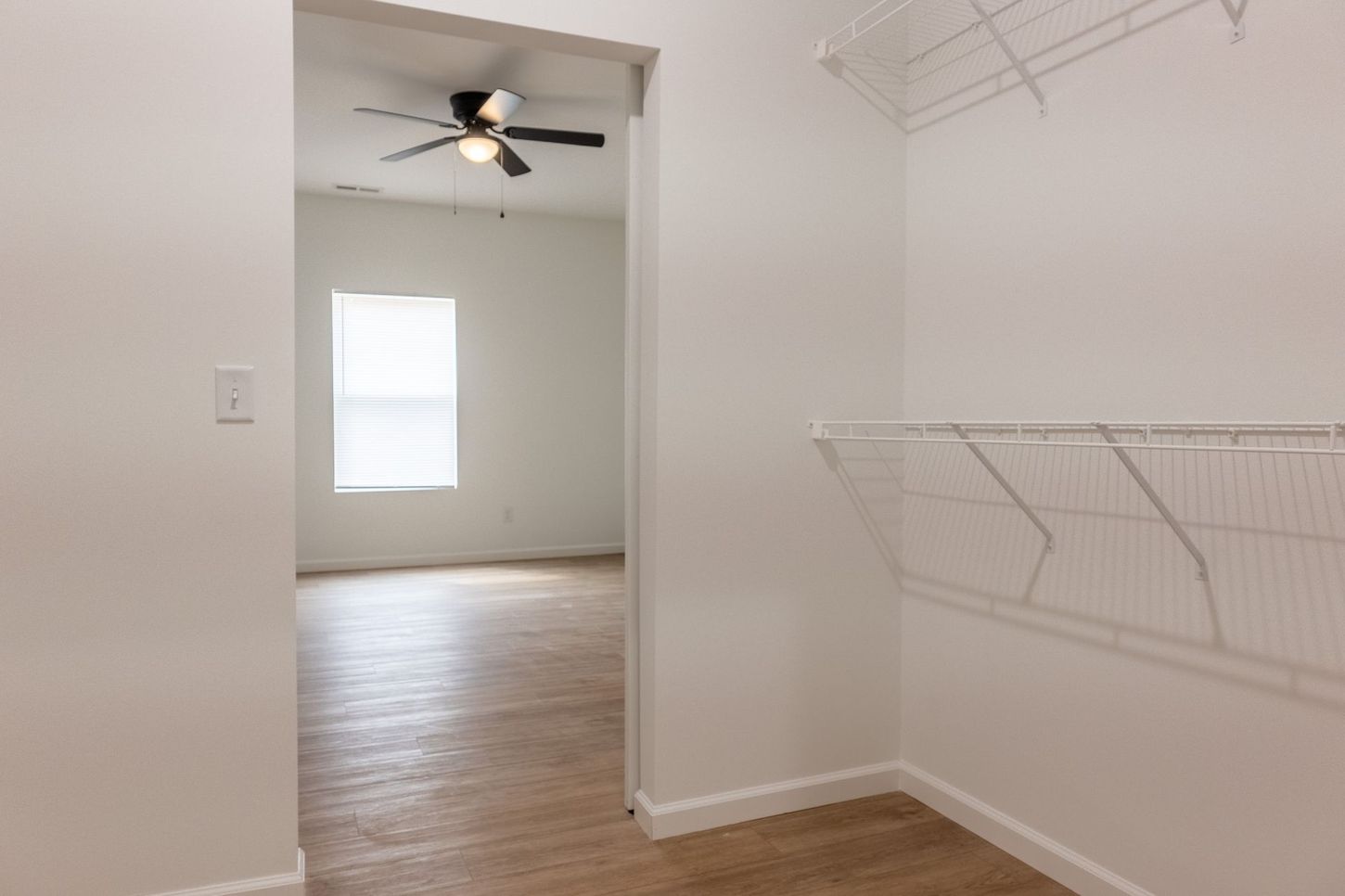 Empty walk-in closet with white wire shelves, wood floors, and a ceiling fan in the adjoining room.