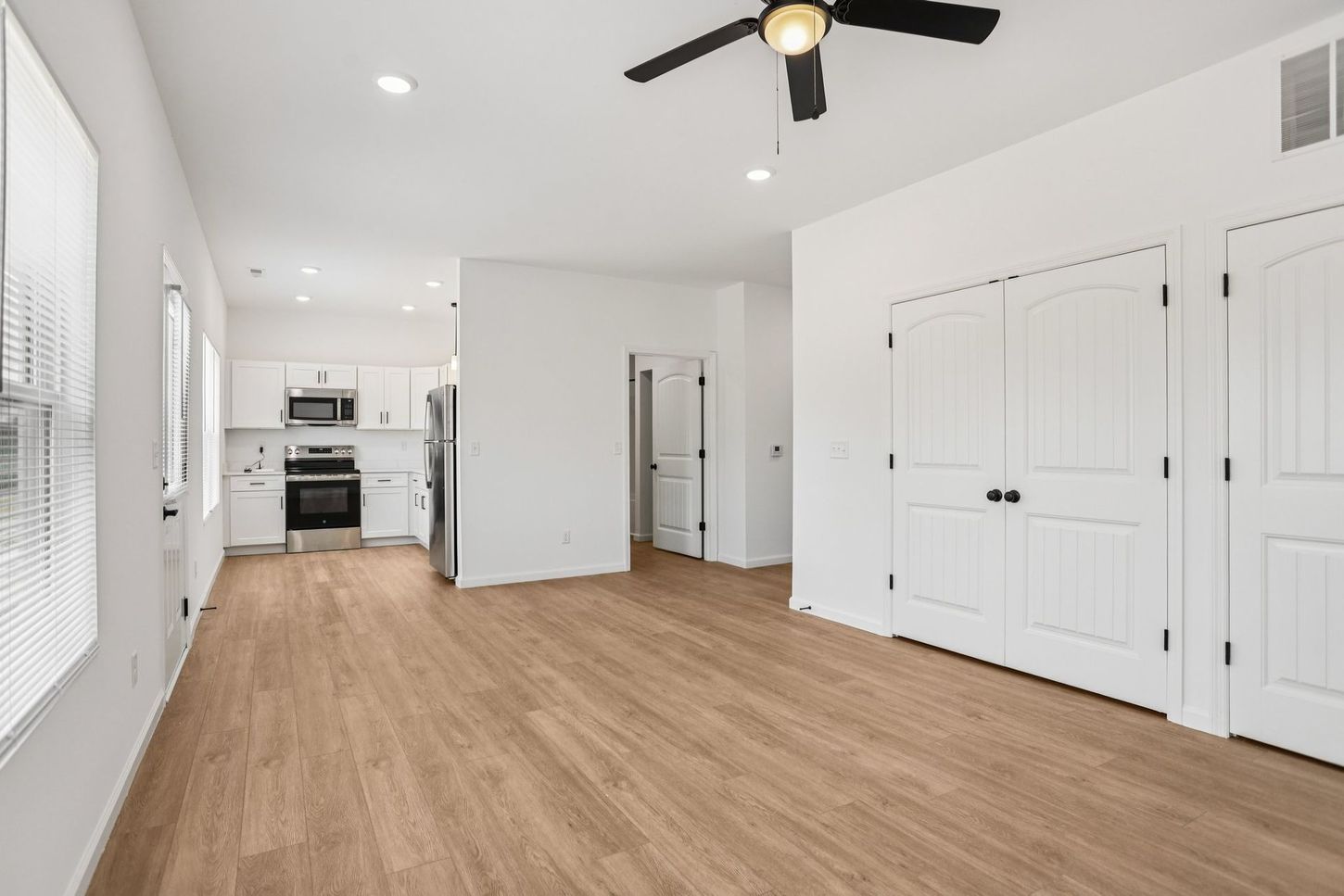 Bright empty room with wood floors, white walls, ceiling fan, and kitchen visible in the back