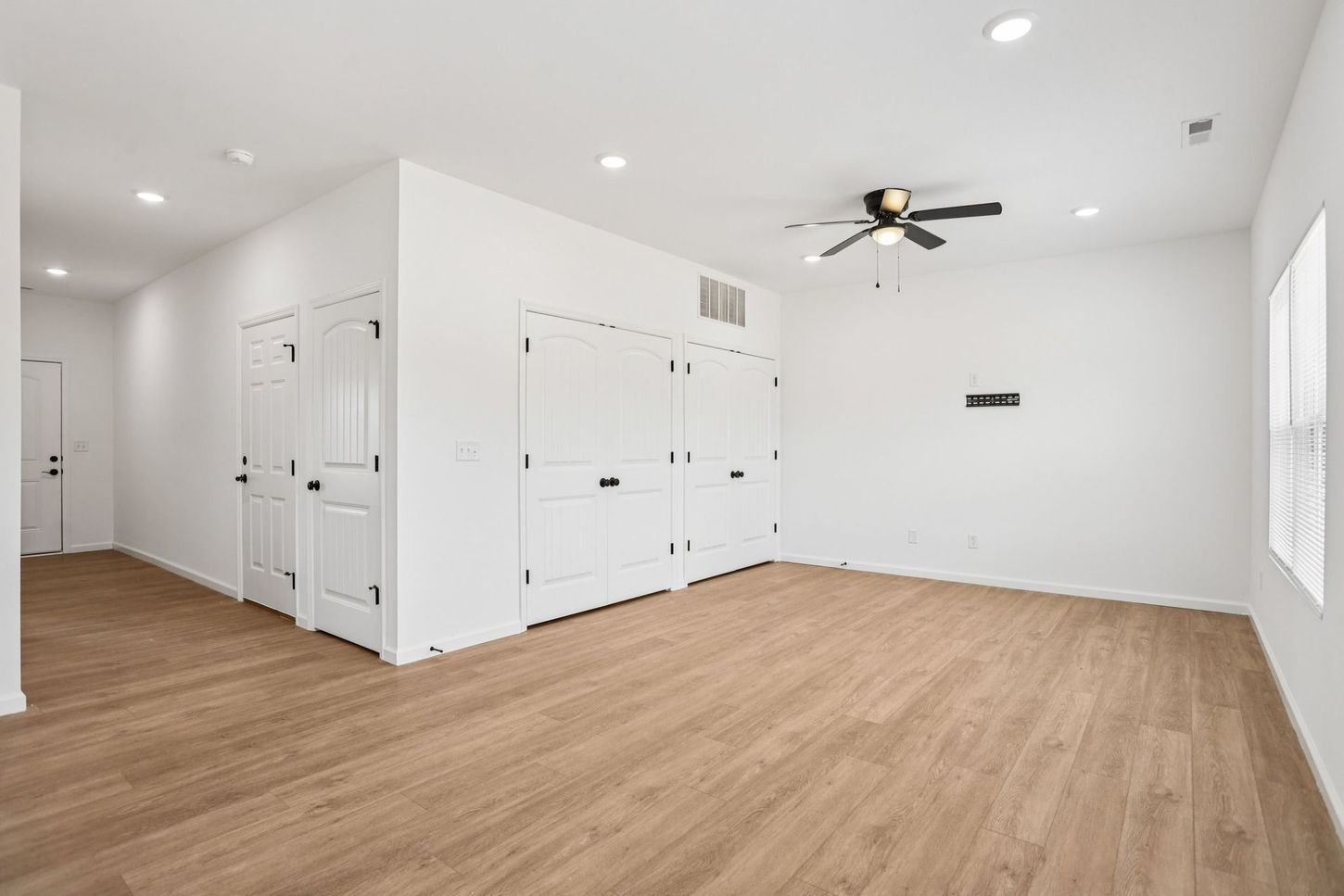 Empty white room with light wood floors, ceiling fan, recessed lights, and multiple doors.
