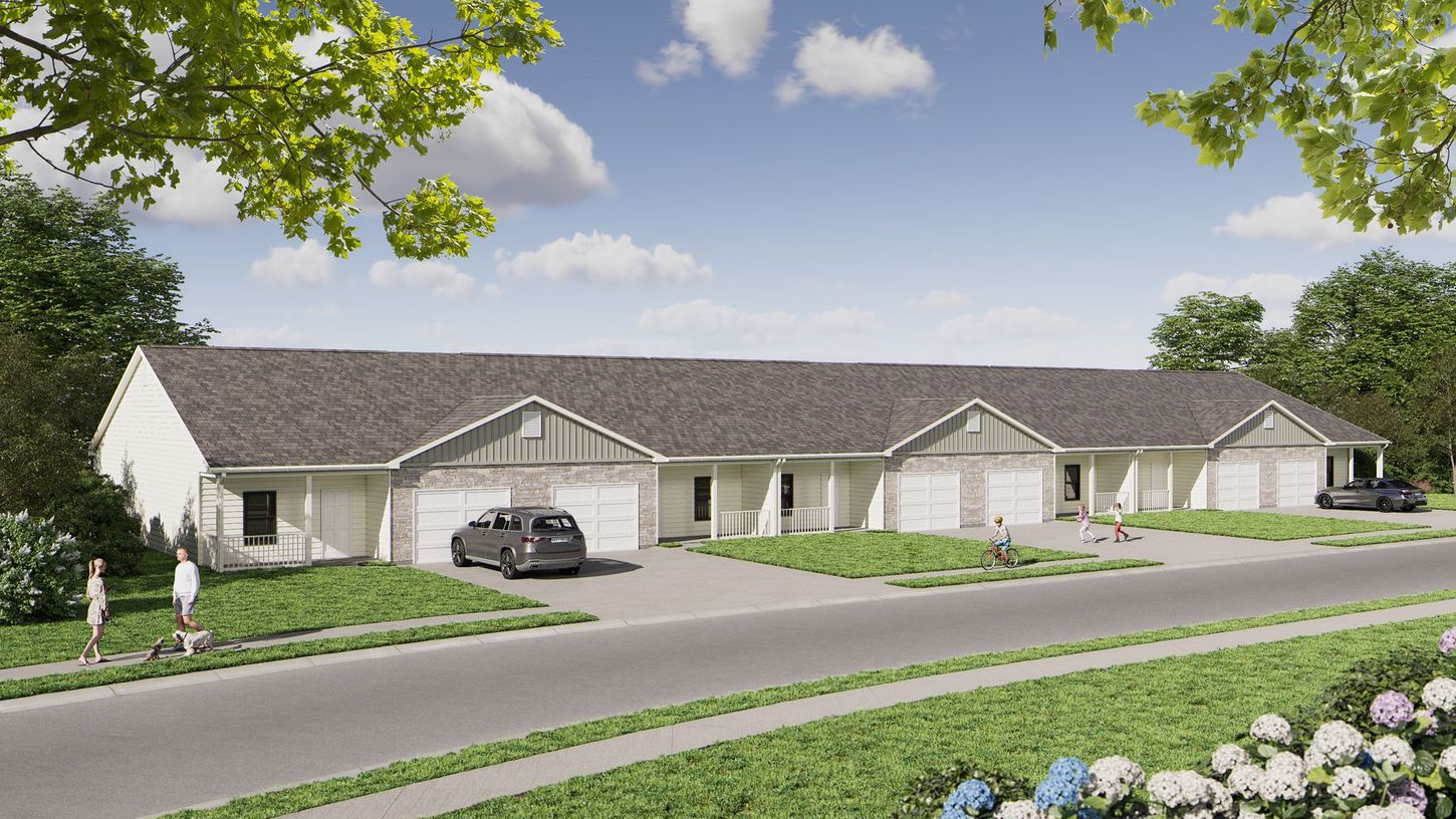 Row of beige townhouses with attached garages beside a road, framed by green trees and flowers