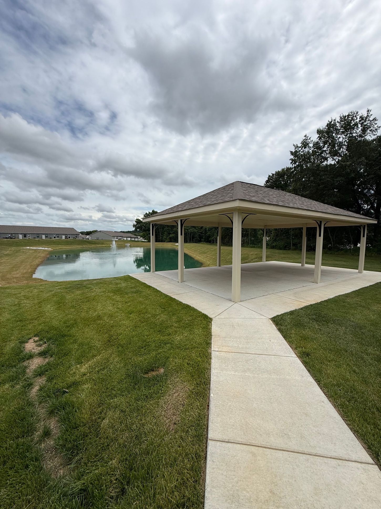 Covered pavilion beside a pond with a concrete path and cloudy sky