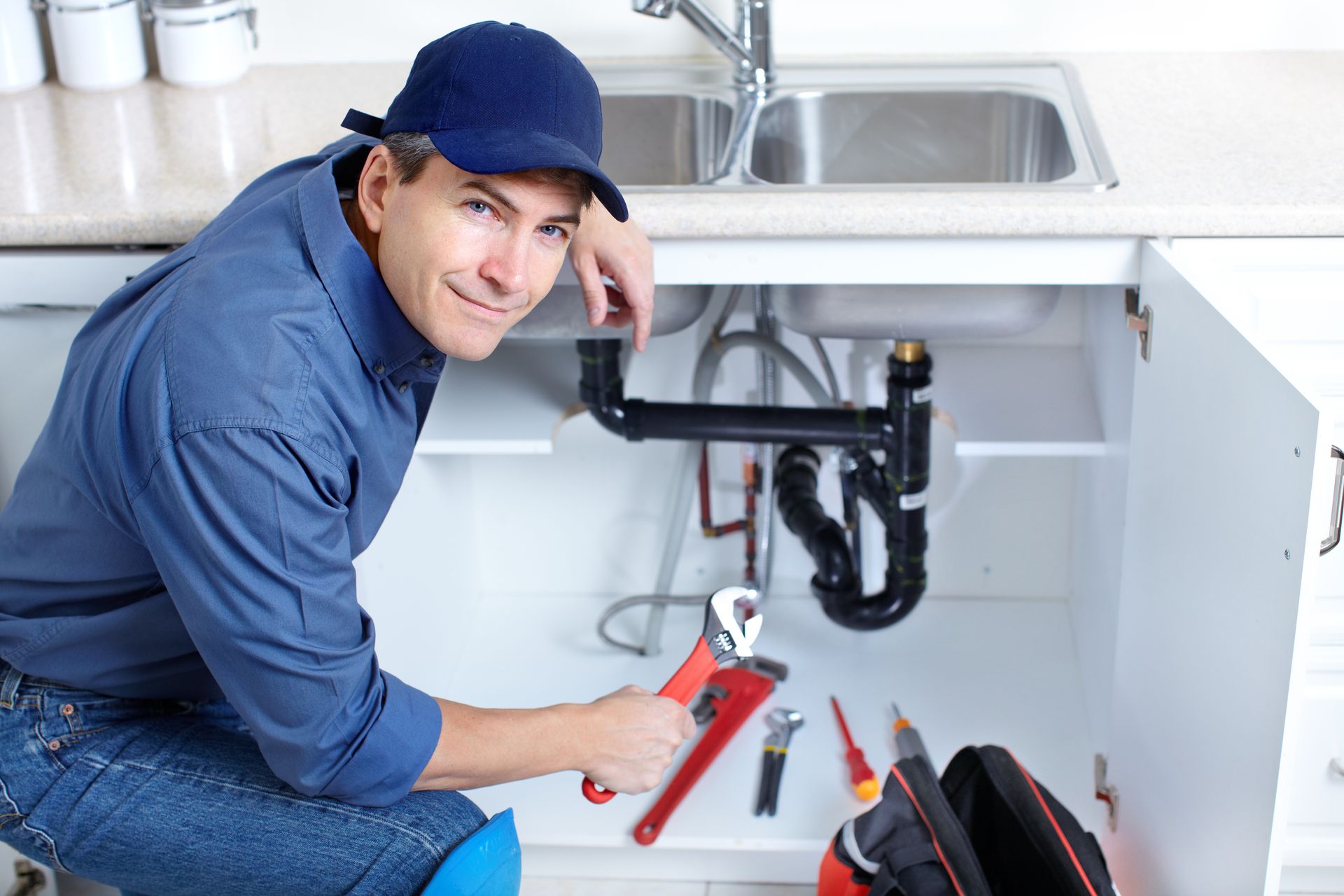Plumber repairing kitchen sink pipes with a wrench under an open cabinet.