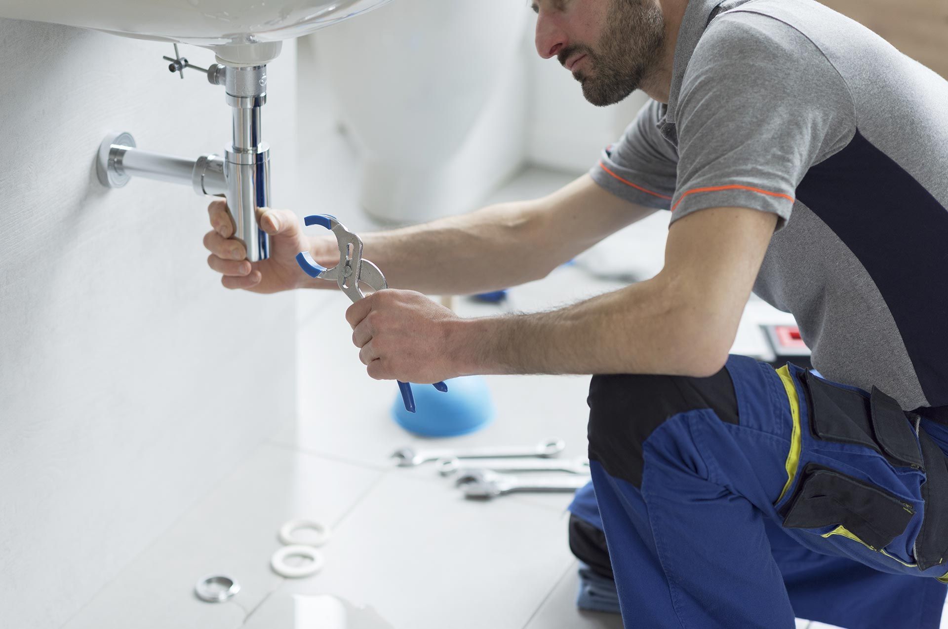 Plumber kneeling, working on sink pipes with pliers, in a bathroom. Plumber kneeling, working on sink pipes with pliers, in a bathroom.