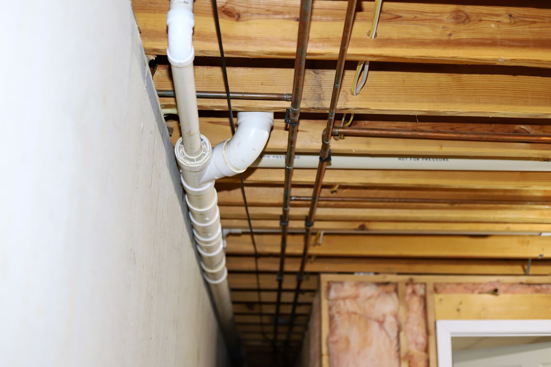 White plumbing pipes and copper pipes run along wooden joists in a basement.