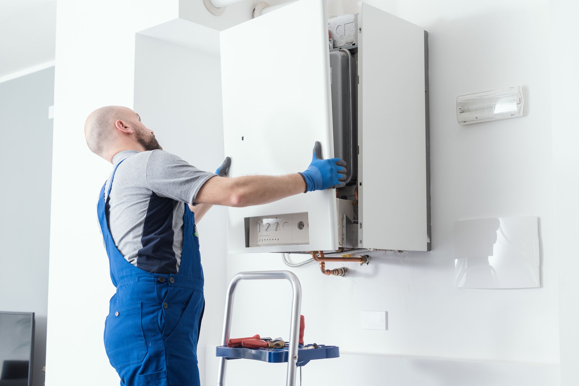 Man in blue overalls installing a white wall-mounted boiler, using a small ladder.