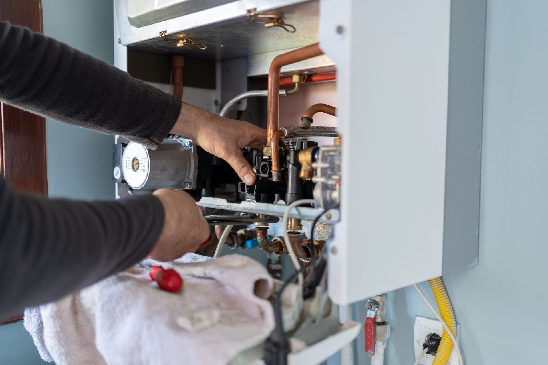 Person working on a heating system. Copper pipes, open white unit. Hands using tools.