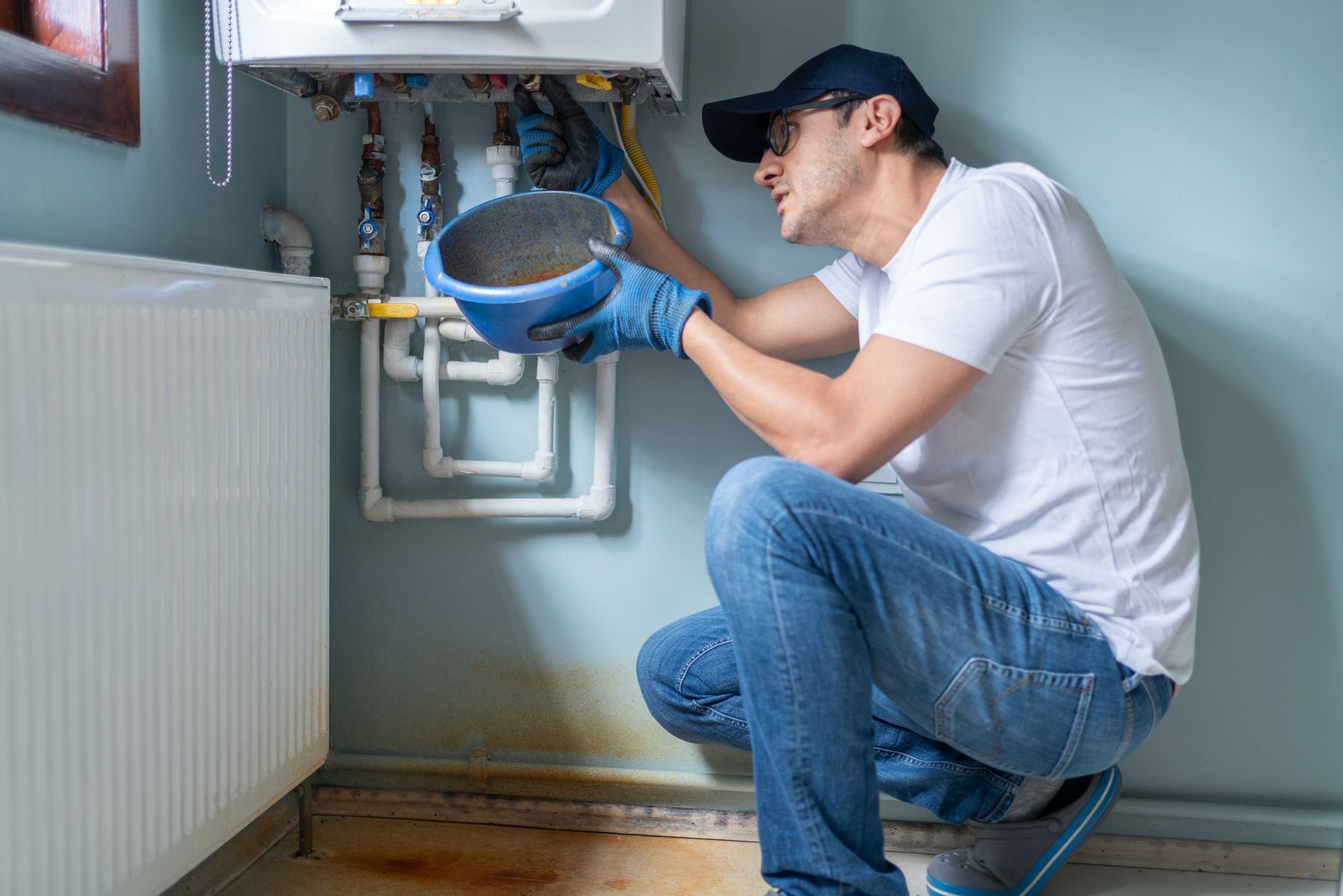 A person in a baseball cap and jeans repairs a boiler, holding a blue bucket.