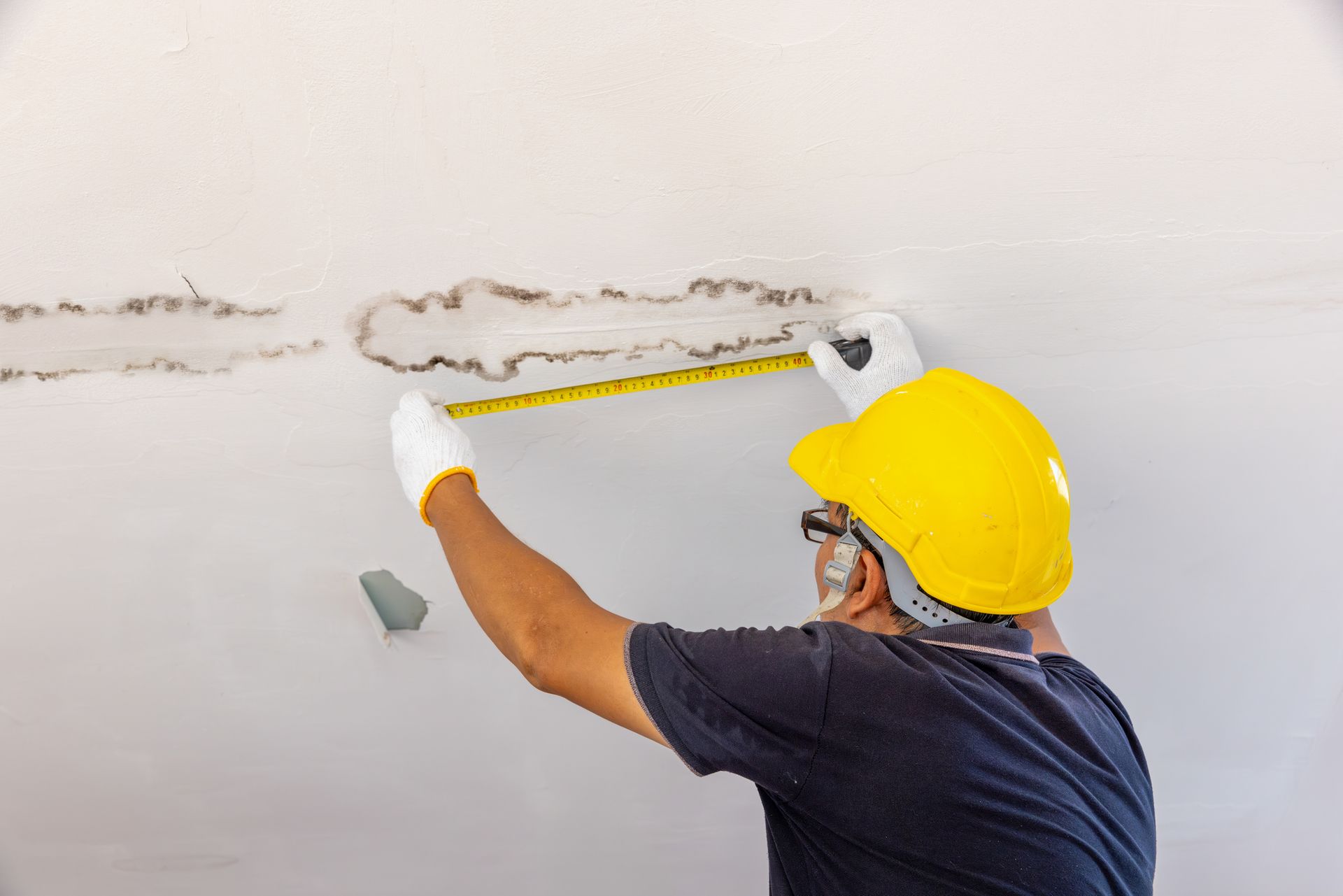 Person in a yellow hard hat measures water damage on a ceiling with a tape measure.