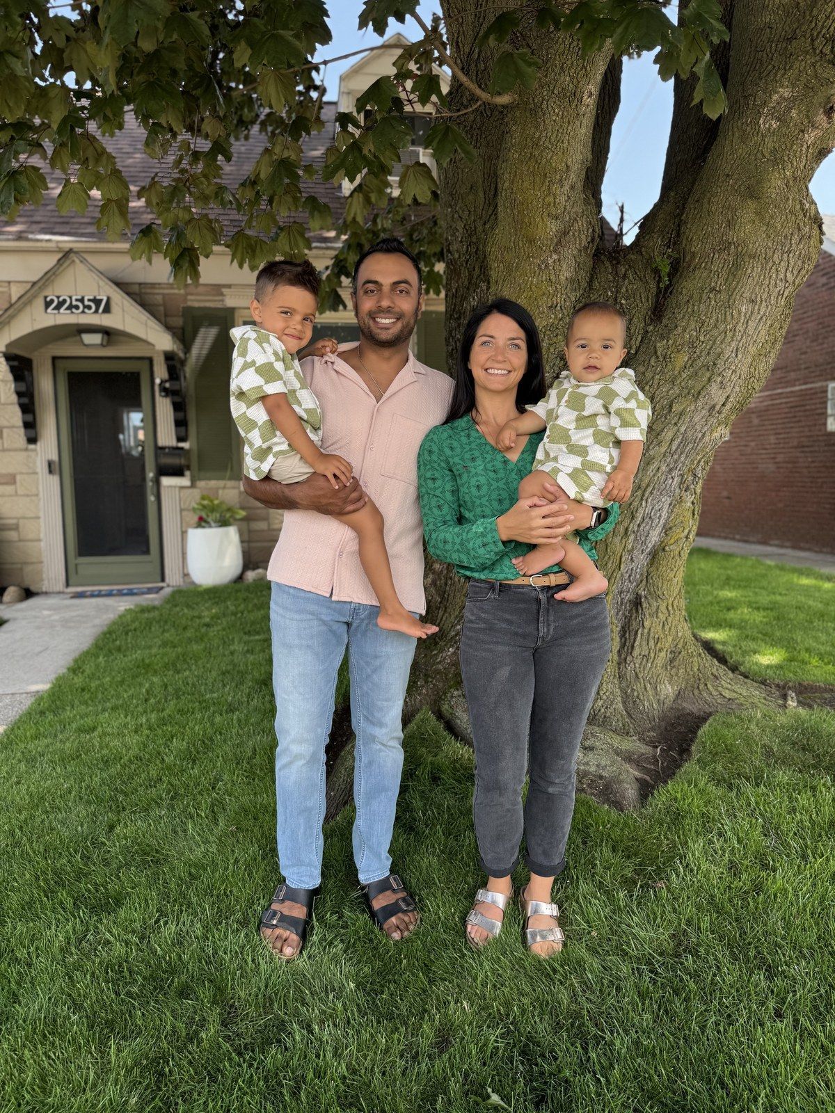 Family of four standing under a large tree in front of a house, smiling and holding two small children.