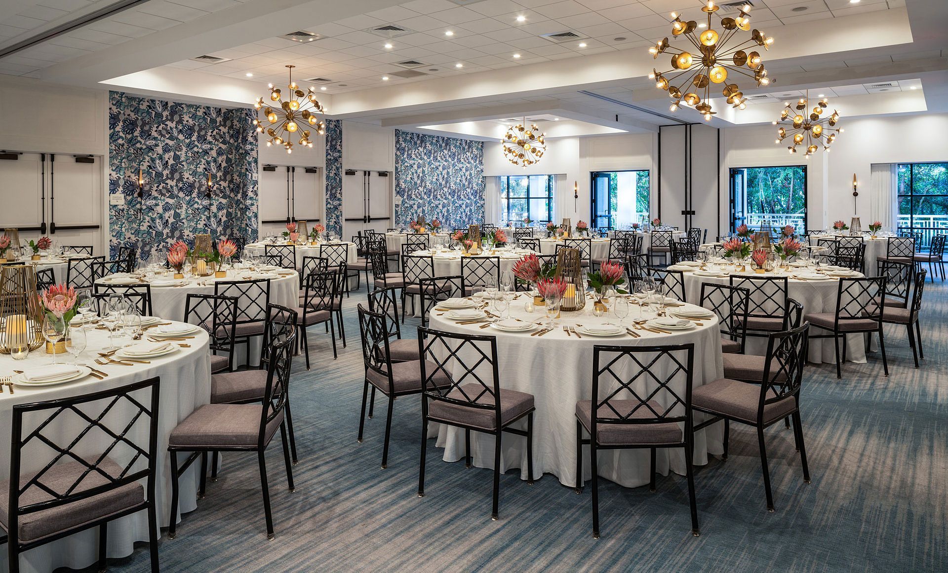 Banquet hall set for event with round tables, chairs, floral centerpieces, decorative walls, and chandeliers.