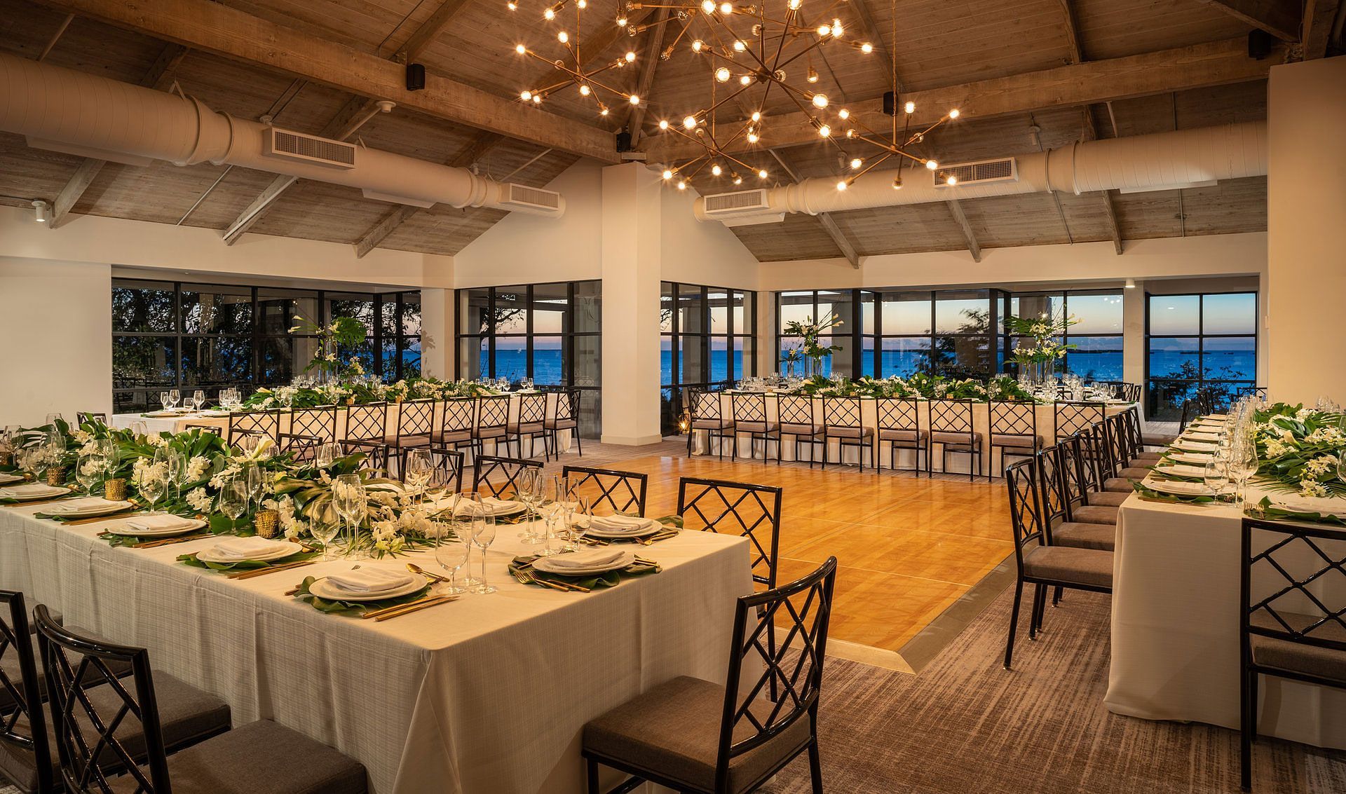 Formal dining room set for an event; tables with floral centerpieces, ocean view through windows.