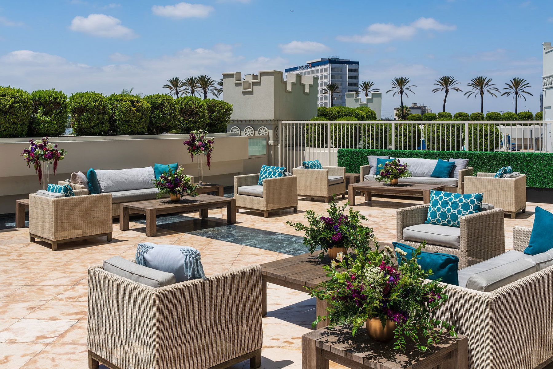 Rooftop lounge with wicker furniture, teal cushions, and floral centerpieces. Overlooks ocean and palm trees.