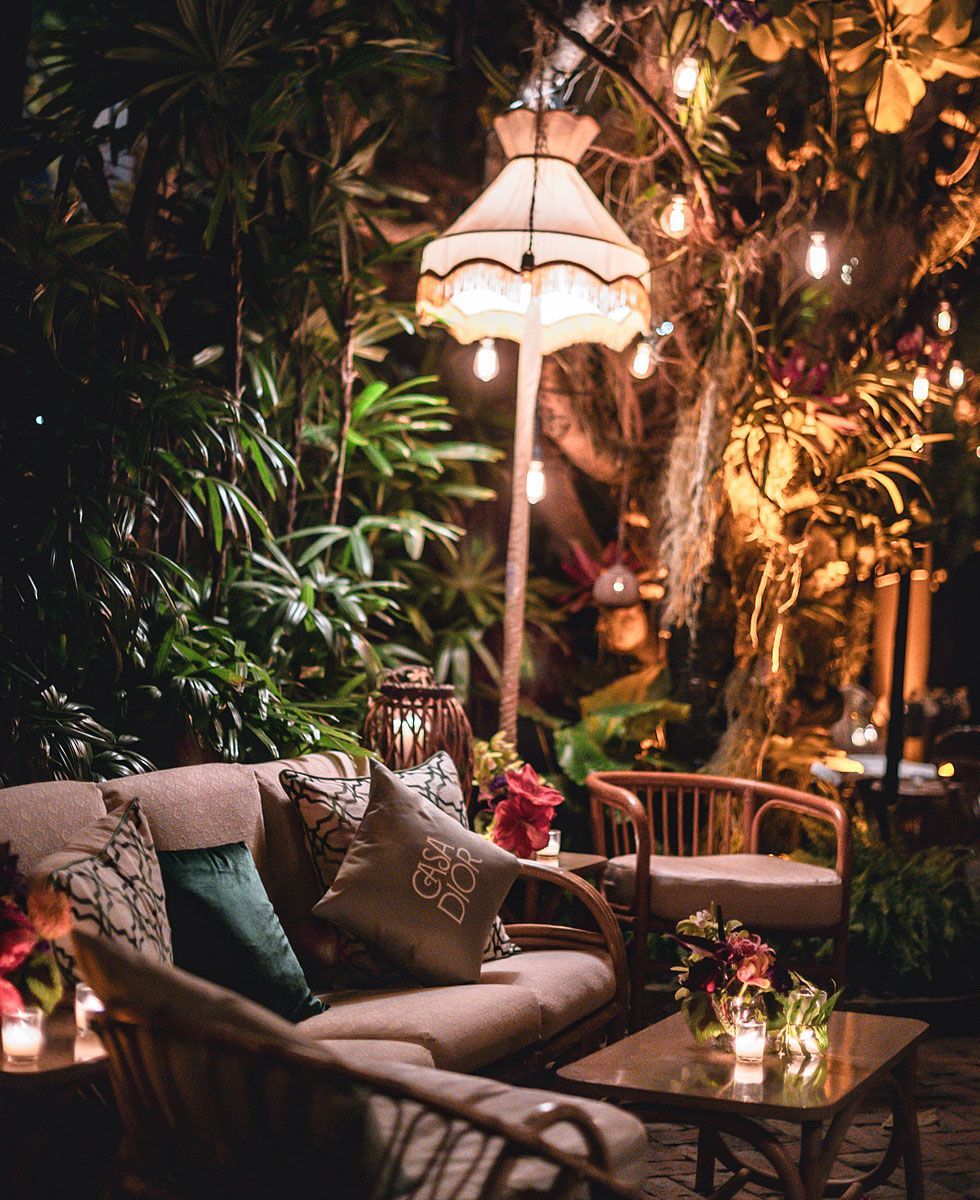 Outdoor lounge with sofa, chair, and table, illuminated by warm lights and surrounded by plants.