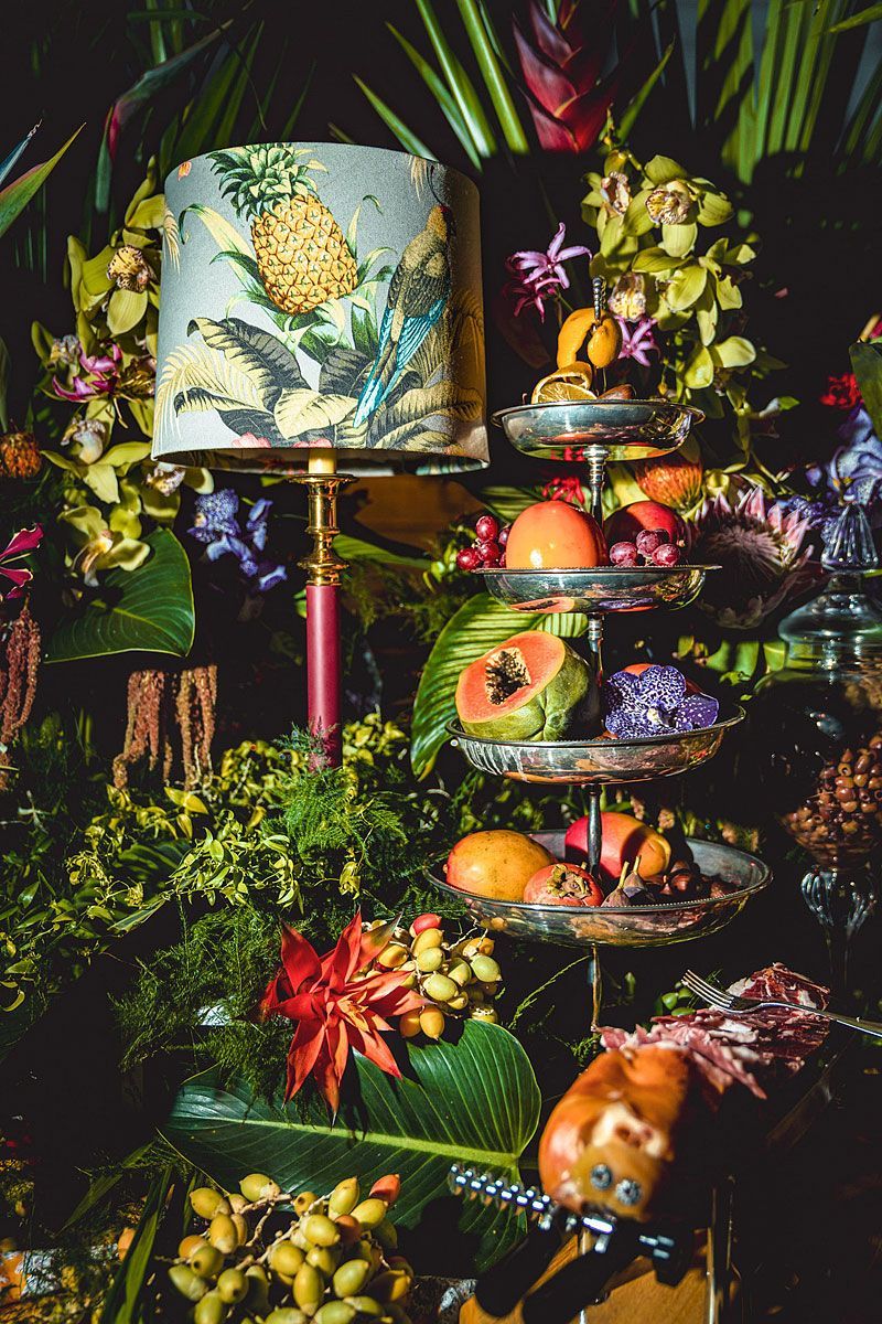 Lamp with floral shade, tiered tray of fruit, surrounded by lush tropical flowers and greenery.