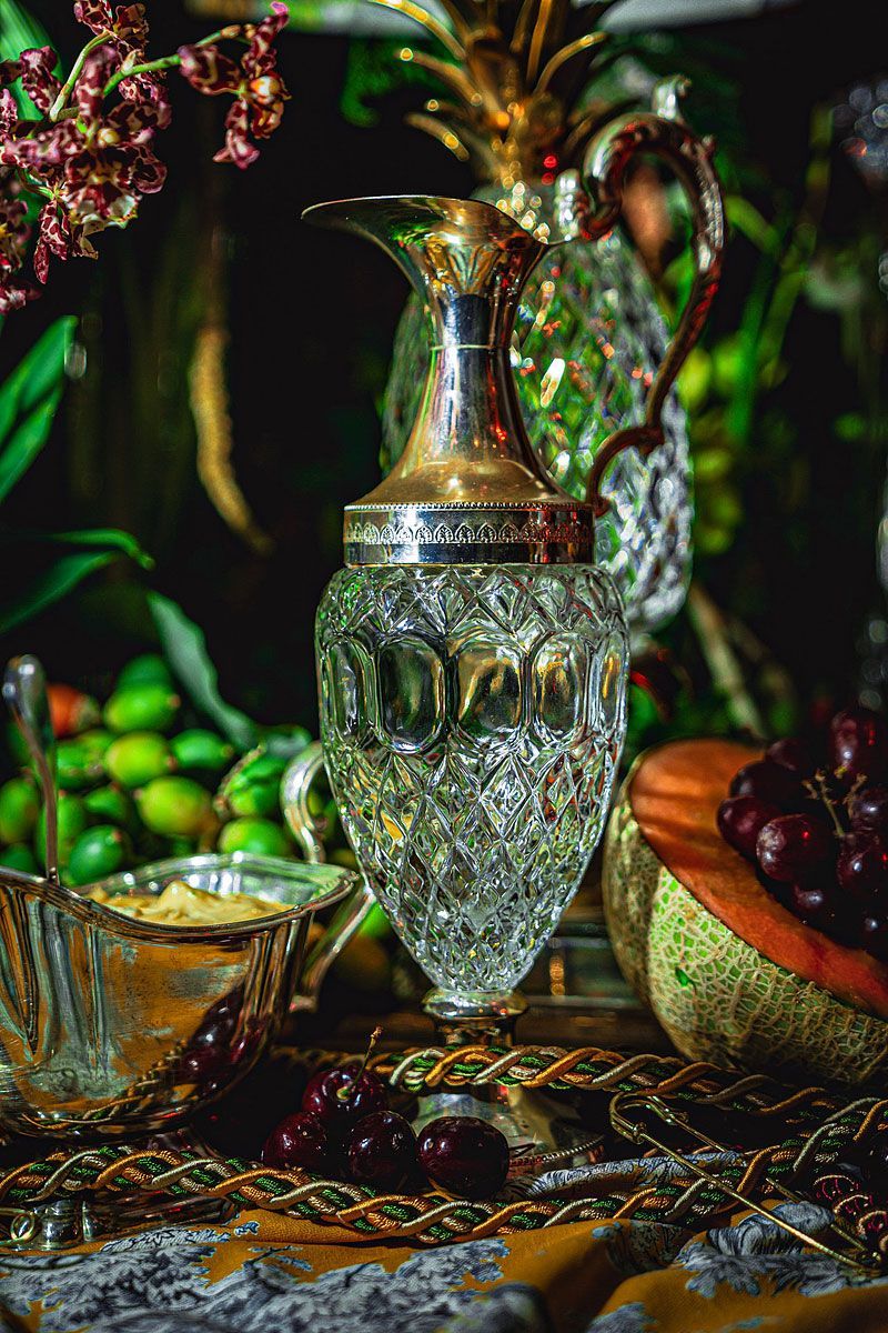 Crystal decanter with silver top, surrounded by fruit, vines, and ornate serving dishes.