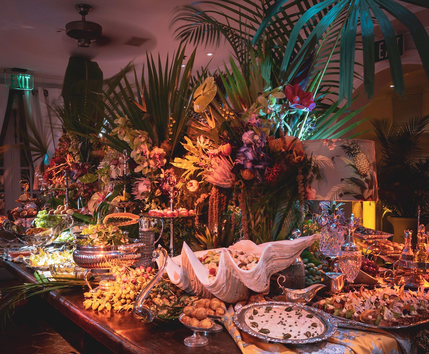 Buffet table with tropical foliage and floral arrangements, food displays, and decorative items.