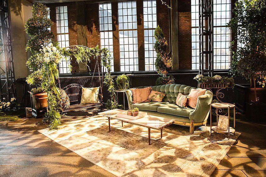 Lounge area with green velvet sofa, vintage accents, and lush greenery in a brick-walled room with large windows.