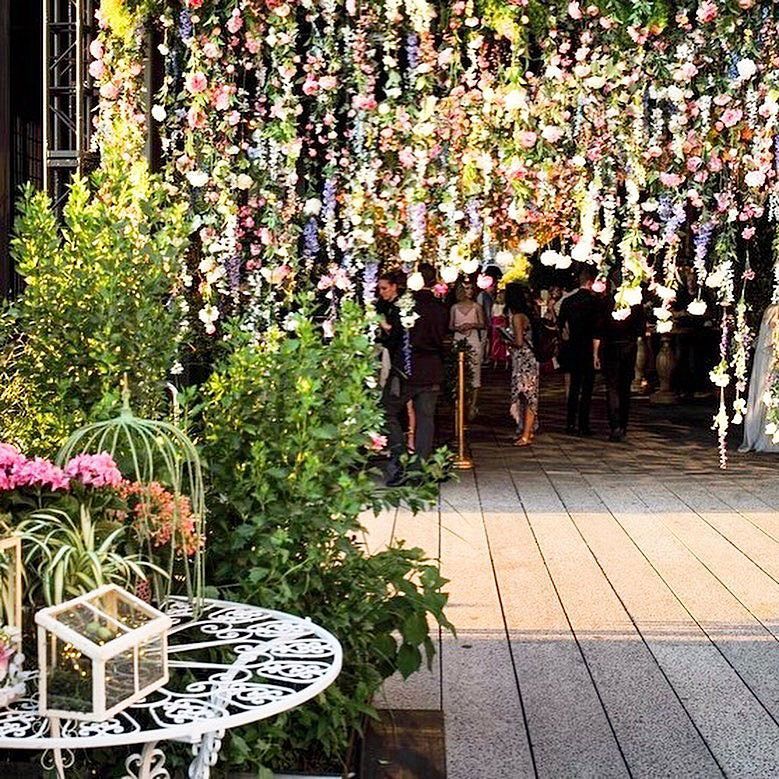 Floral archway with hanging flowers; people walking beneath. White table with decorative plants in foreground.