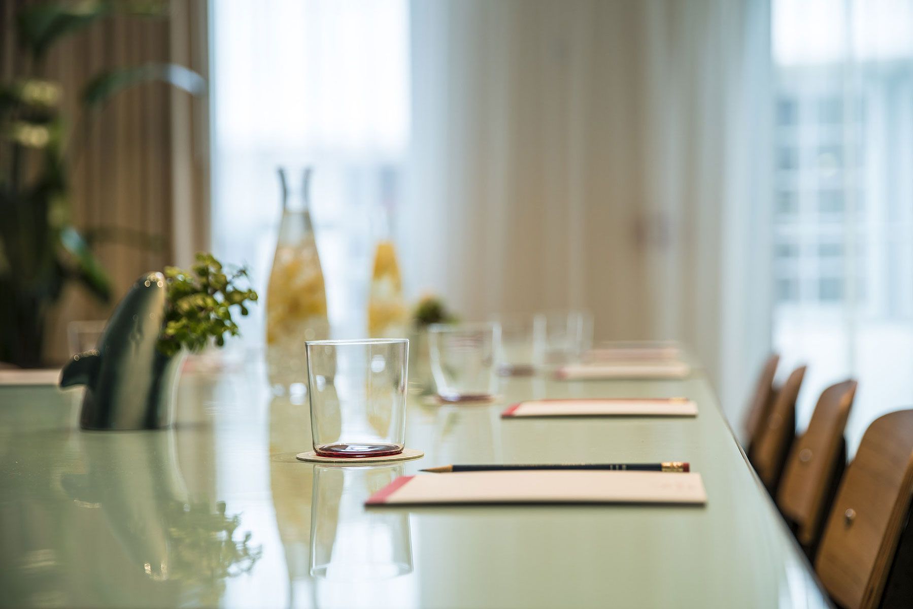 Long, glass table set for a meeting; water pitcher, glasses, notepads, and chairs arranged.