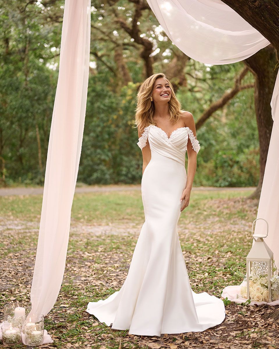 Woman in white off-shoulder wedding dress smiles outdoors under sheer fabric.