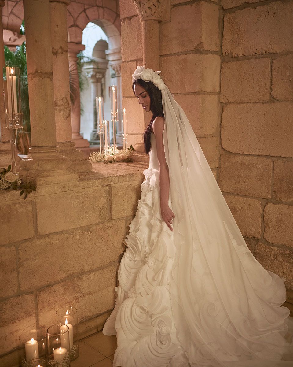 Bride in a white gown and veil stands near a stone wall with candles.