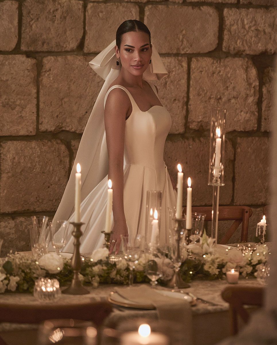 Woman in wedding dress stands near a table with candles, stone wall backdrop.