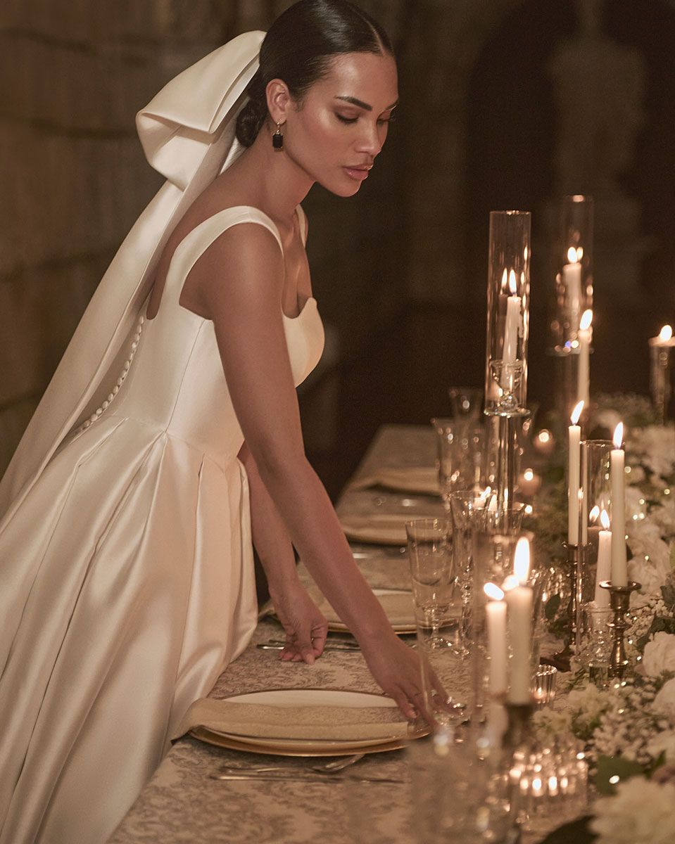 Woman in satin wedding dress arranging place setting at candlelit table.
