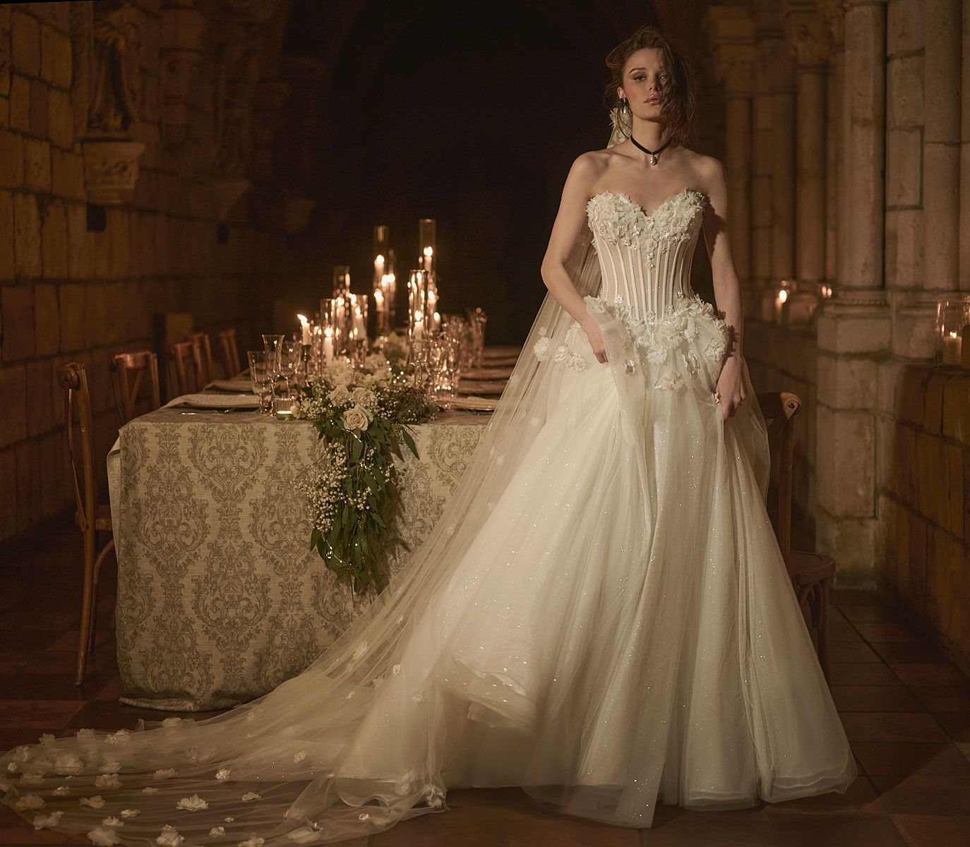 Woman in white wedding gown with long train, posed in front of a candlelit table, stone interior.