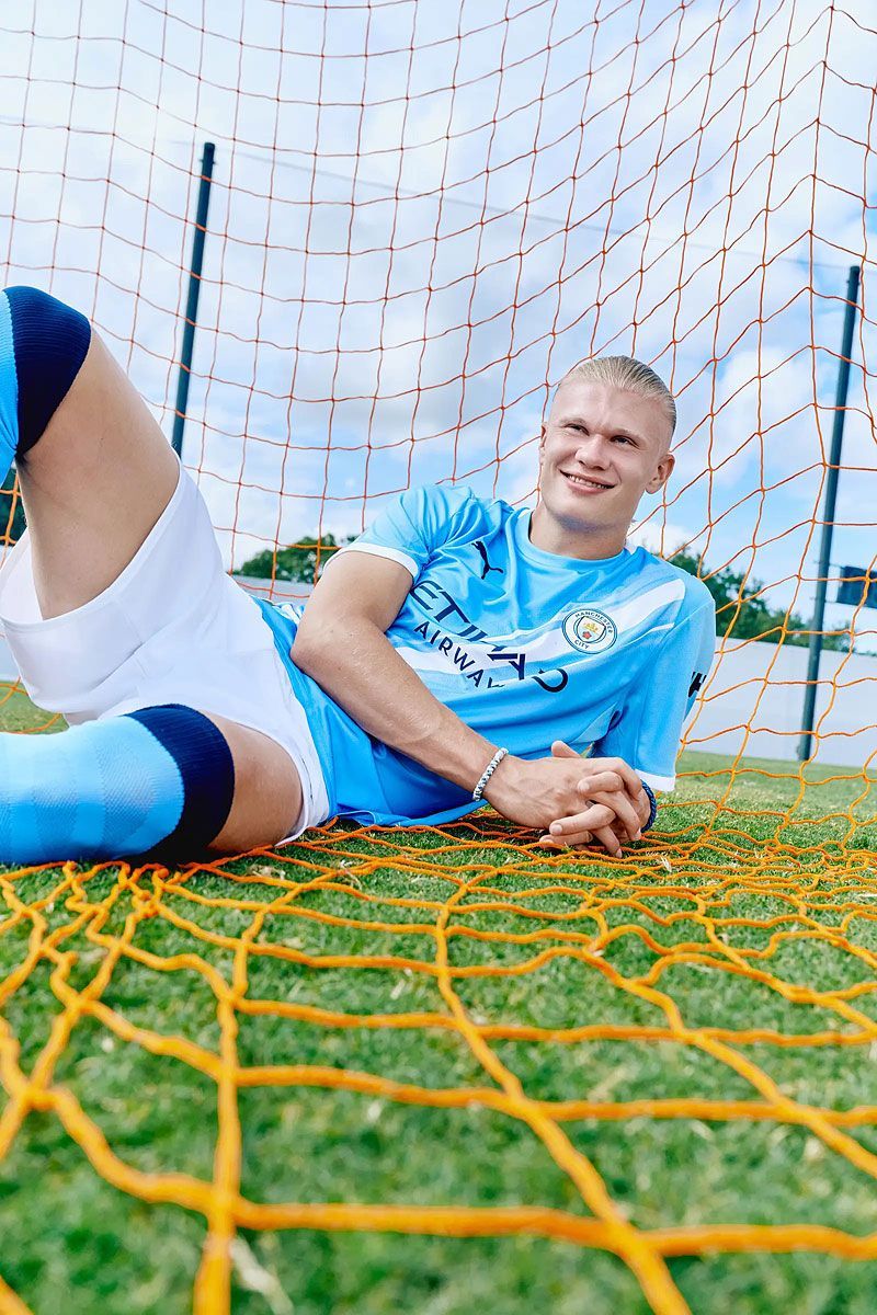 Soccer player Erling Haaland in a sky blue jersey smiles, lying on a green field behind a goal net.
