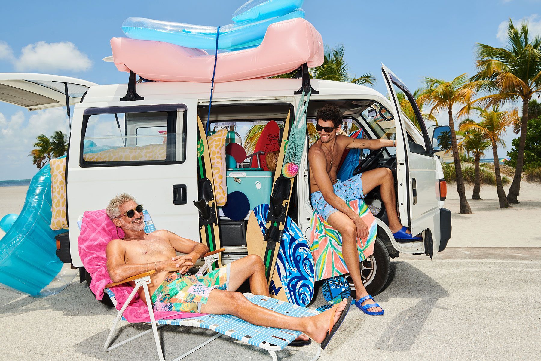 Two men relax by a van packed for the beach with inflatables, surfboards, and palm trees.